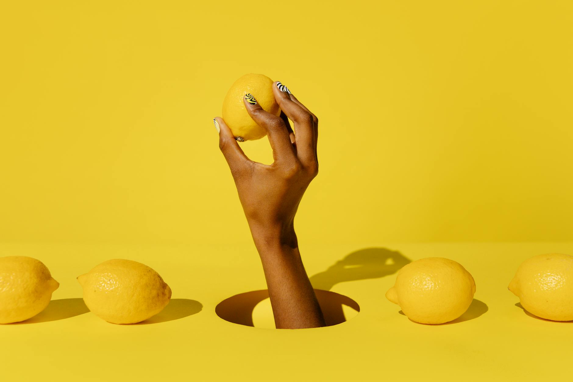 A hand holding a fresh lemon against a vivid yellow background, representing the citrus-inspired design of lemon vibrators