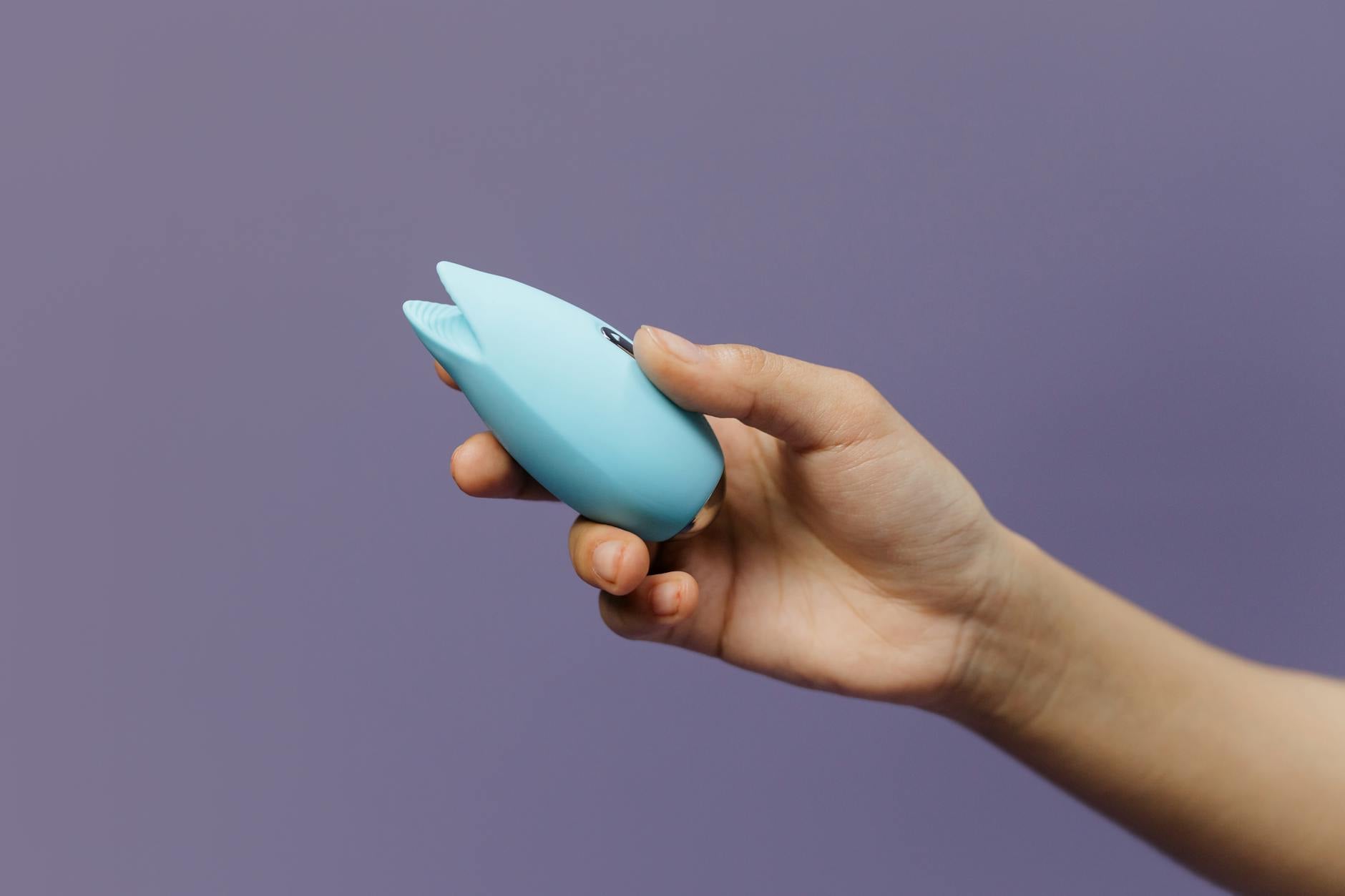 A blue silicone clitoral vibrator held gently in hand against a calm purple background.